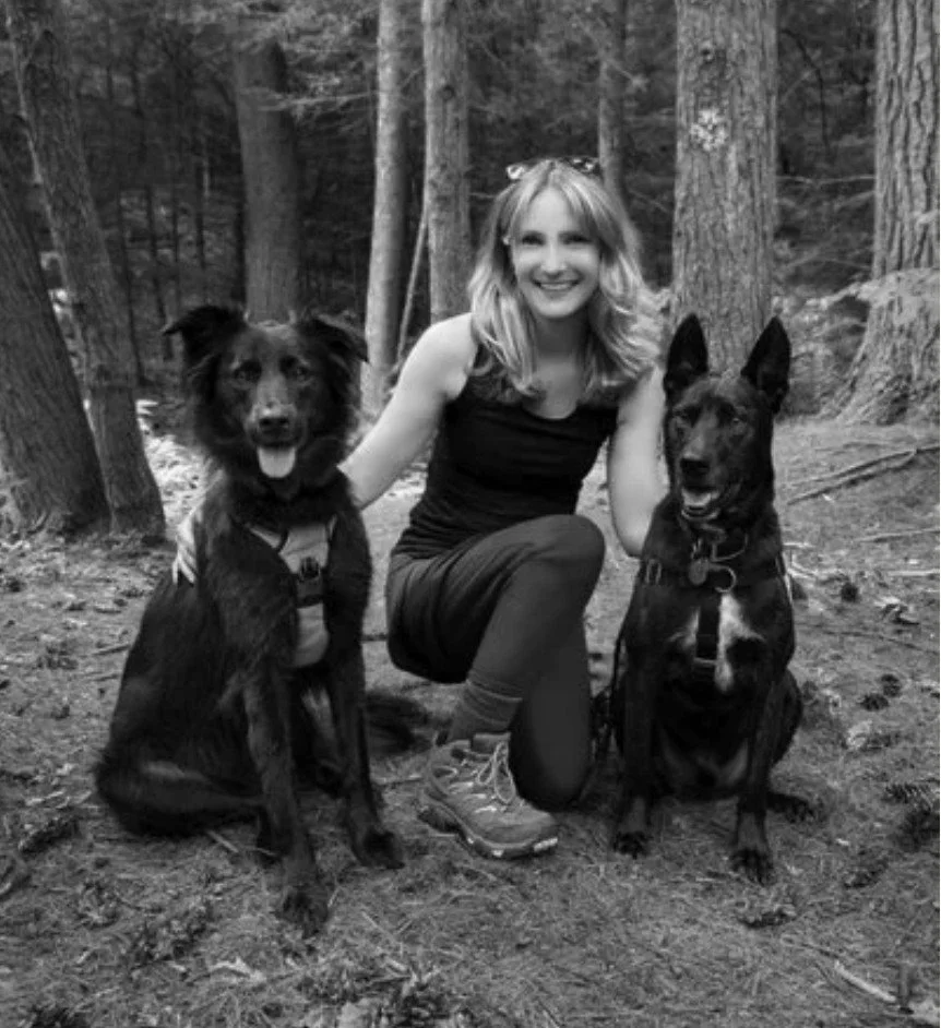 A smiling young woman with blond hair, wearing a sleeveless shirt and hiking boots, kneels on a forest trail with two medium-sized dogs, one on each side. The setting is wooded with tall trees.