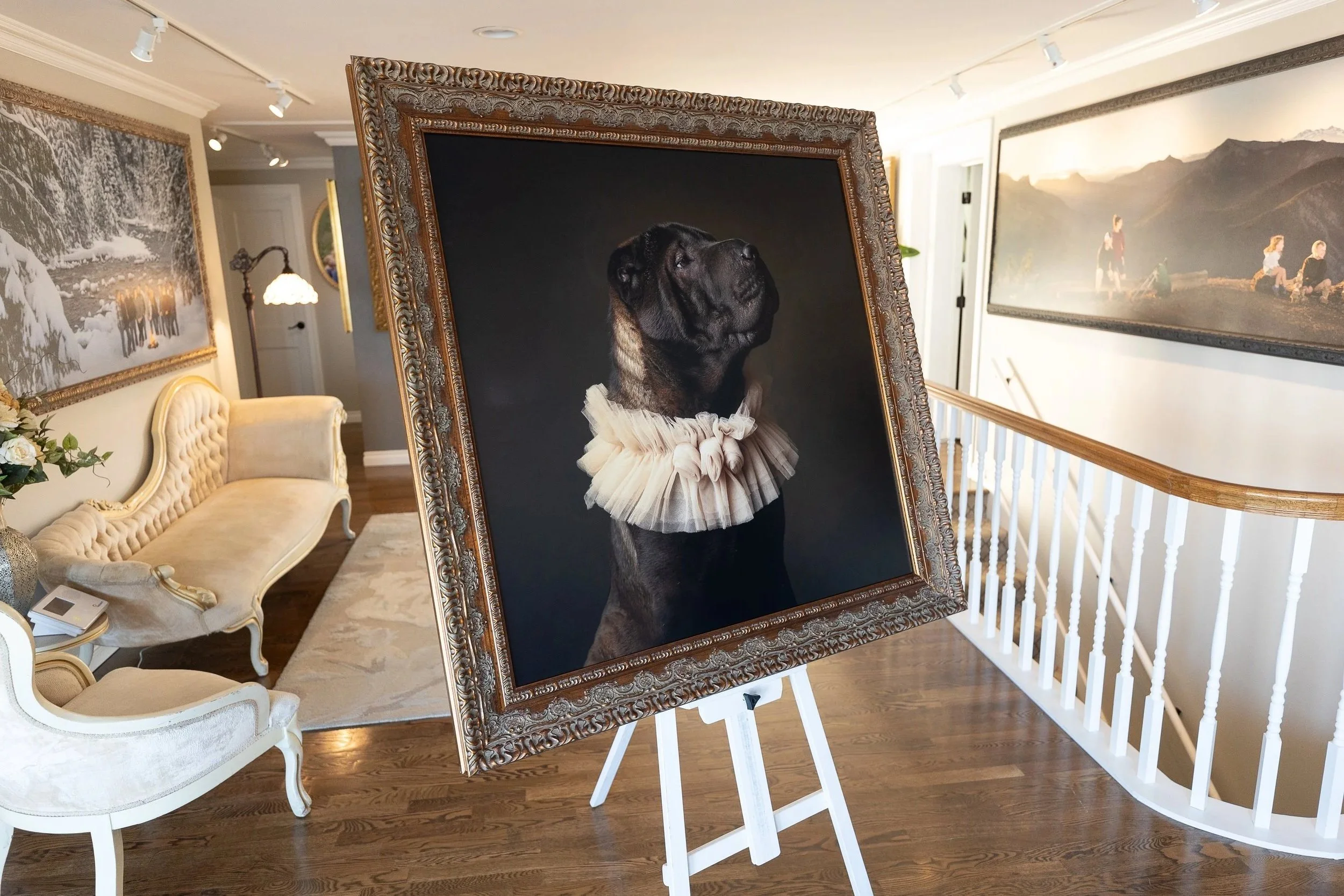 A black dog with a ruffled collar is framed in an ornate gold picture frame, standing on an easel in a well-lit art gallery. The gallery has light-colored walls with various landscape paintings and a cream-colored vintage sofa nearby.