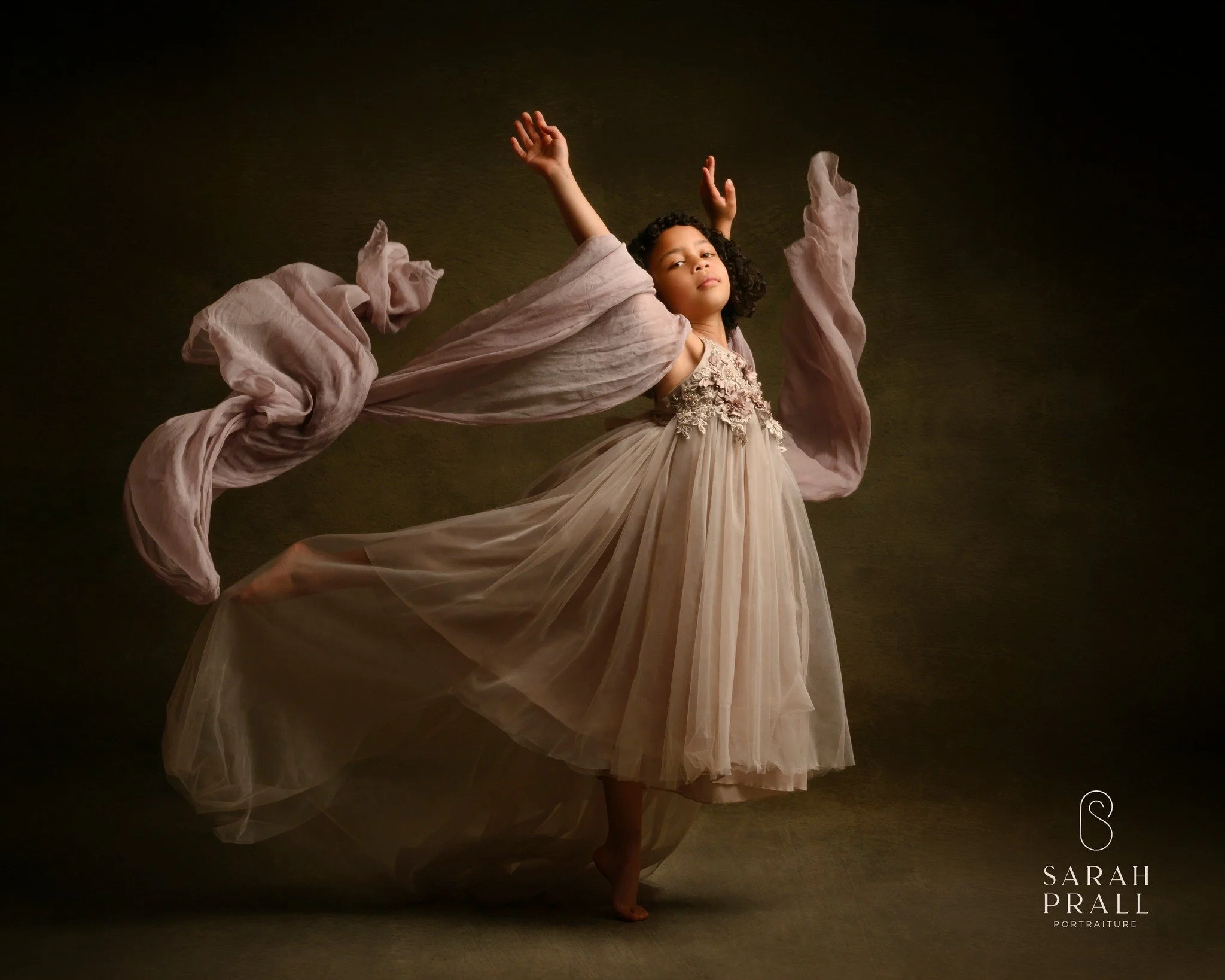 A young girl in a flowing, pale pink dress with lace and floral details performs a ballet dance with her arms raised and one leg lifted, set against a dark background.
