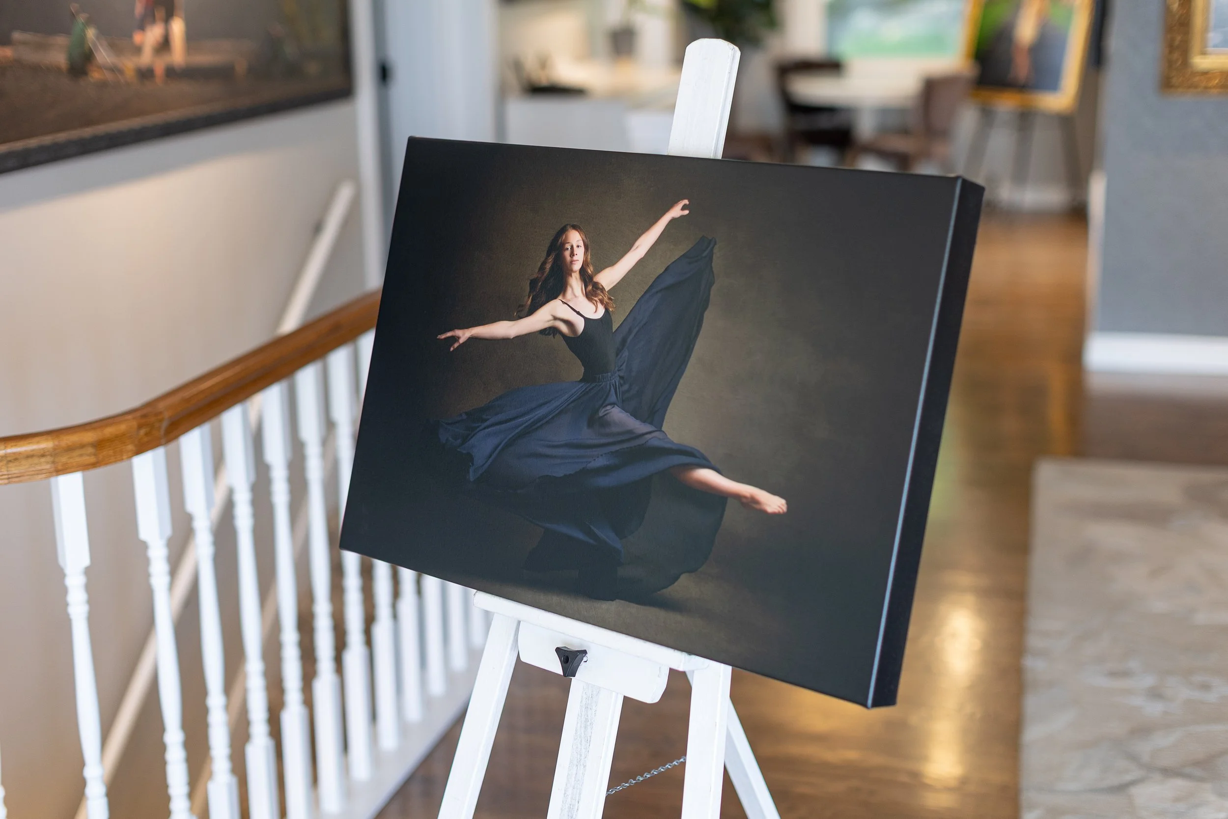 A photograph of a ballerina mid-leap, wearing a black dress with a flowing skirt, displayed on a canvas on an easel inside an art gallery.