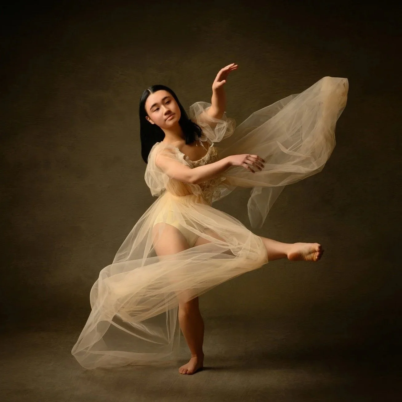 A woman with long black hair dancing in a flowing, sheer beige dress against a dark background.