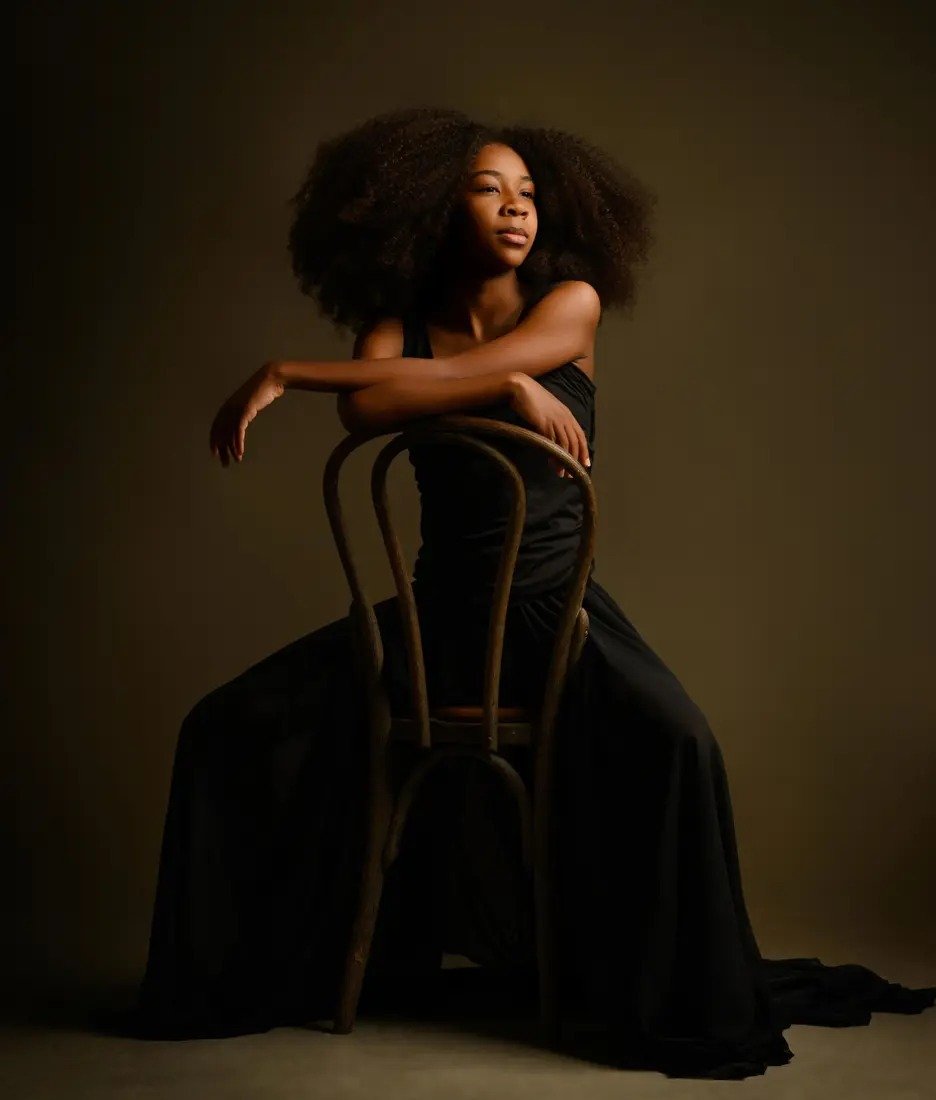 A child in a black dress seated backward on a wooden chair in a fine art portrait