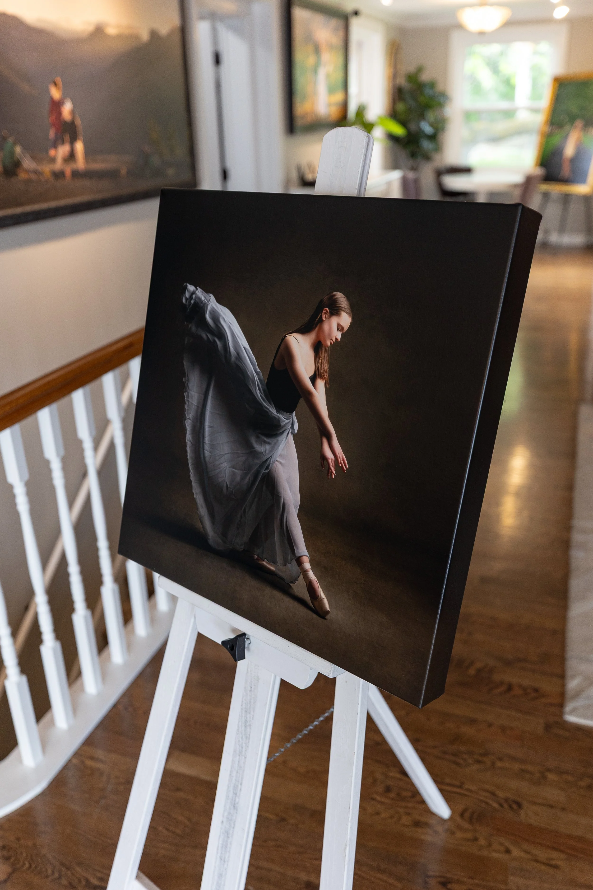 Photograph of a painting depicting a ballerina in a black leotard and gray tutu, standing on pointe with arms extended gracefully. The painting is on an easel, in an art gallery setting with a wooden floor, white walls, and large windows in the background, along with other artwork and a potted plant.