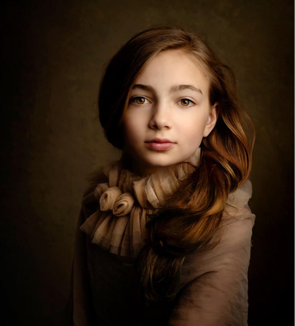 Portrait of a young girl with long reddish-brown hair, wearing a beige outfit with a ruffled collar, against a dark brown background.