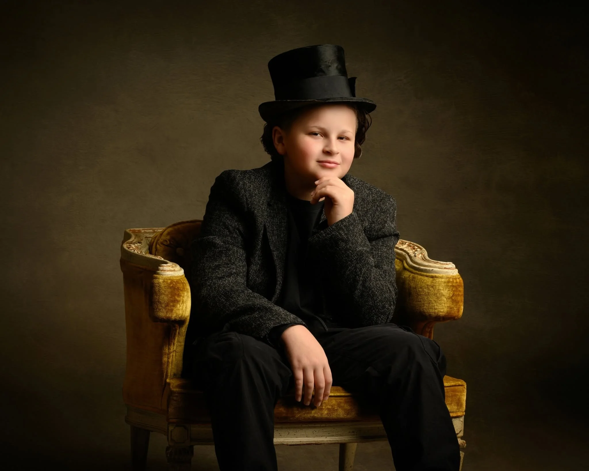 A young boy with a thoughtful expression, wearing a black top hat and a dark blazer, sitting on an ornate yellow vintage chair against a neutral dark background.