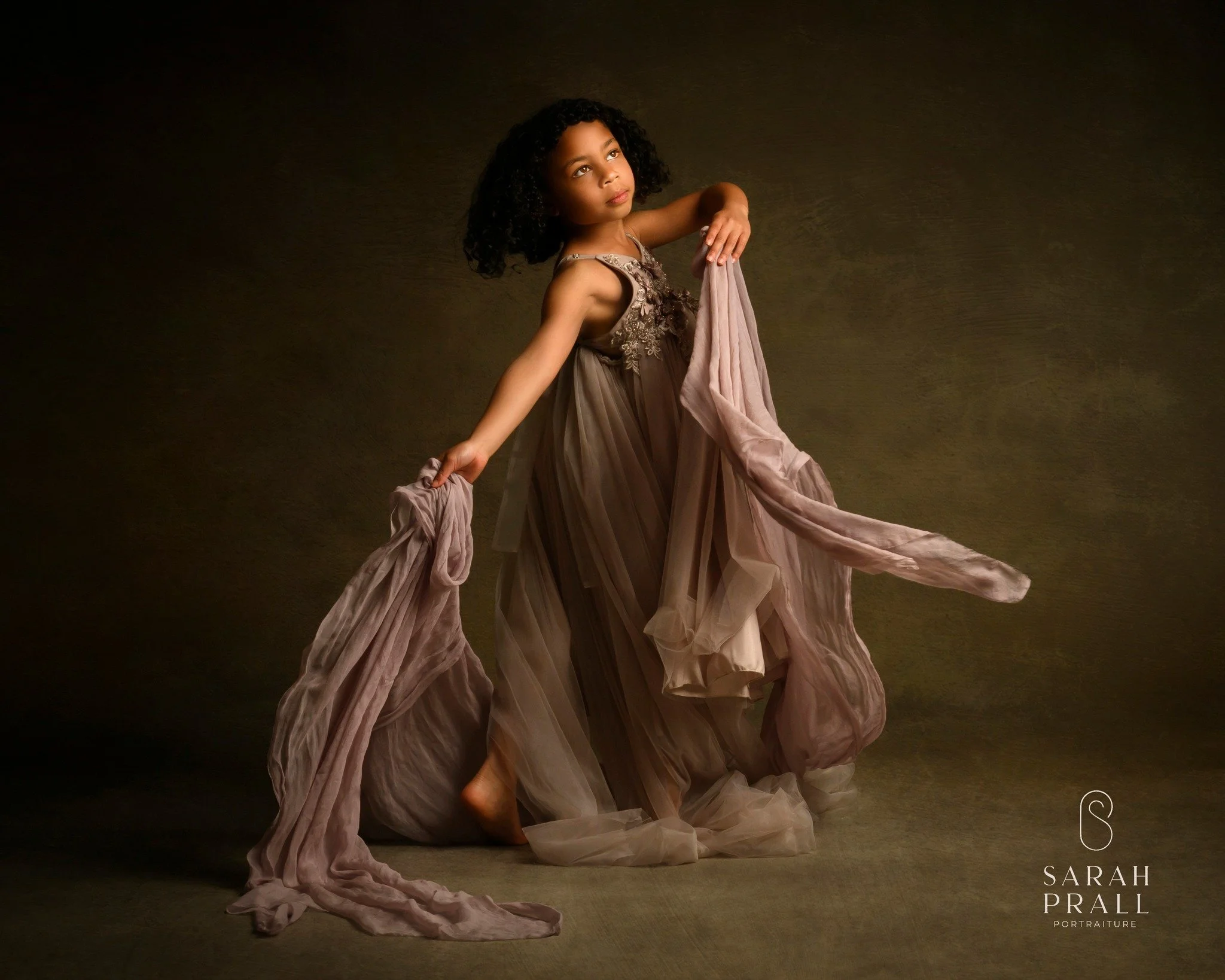Young girl with curly hair in a flowing beige dress standing against a dark background, holding fabric, with a thoughtful expression.