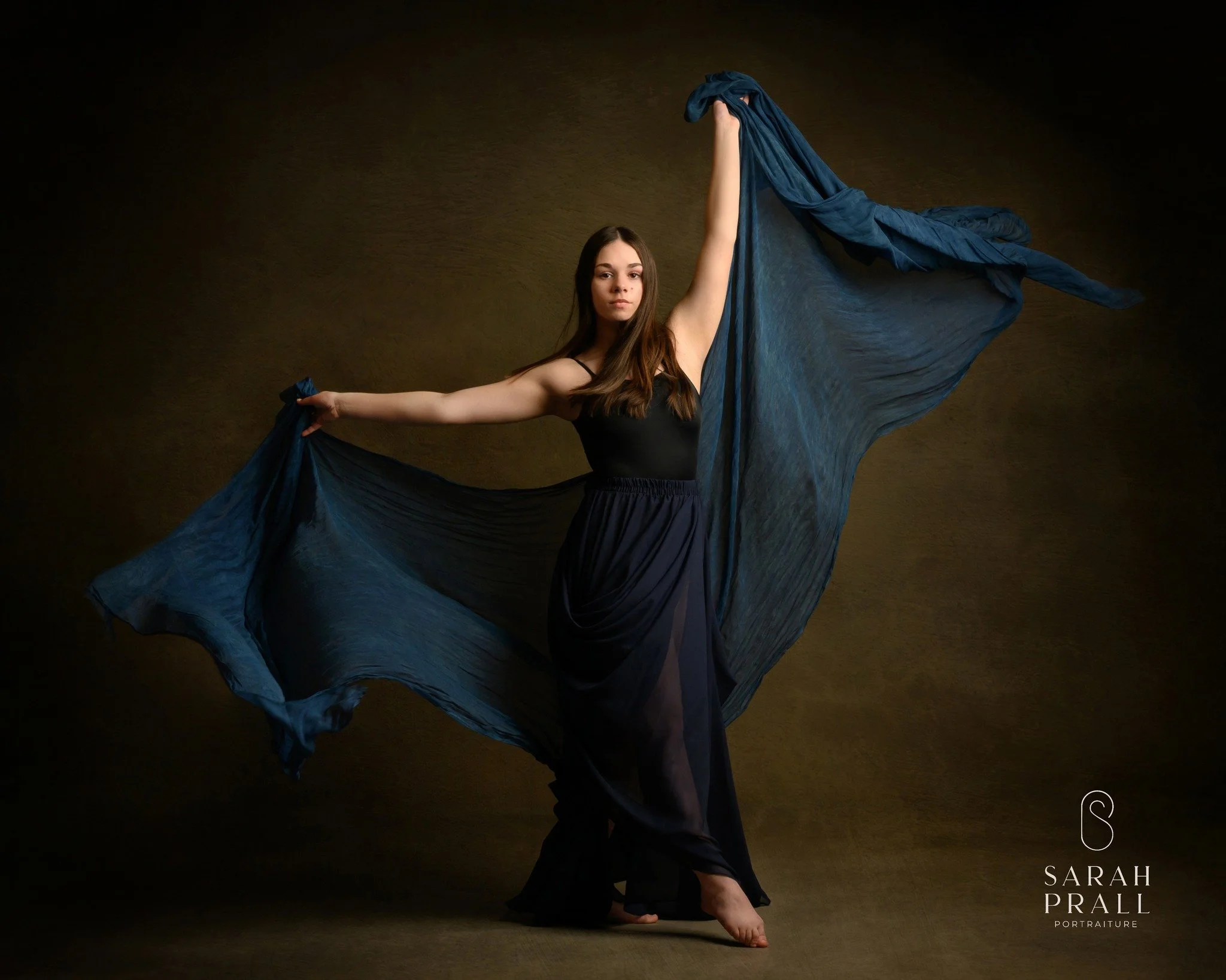 A young woman with long brown hair is posing against a dark background, holding blue fabric that flows around her. She is standing on one foot with her arms extended, wearing a black top and blue skirt.
