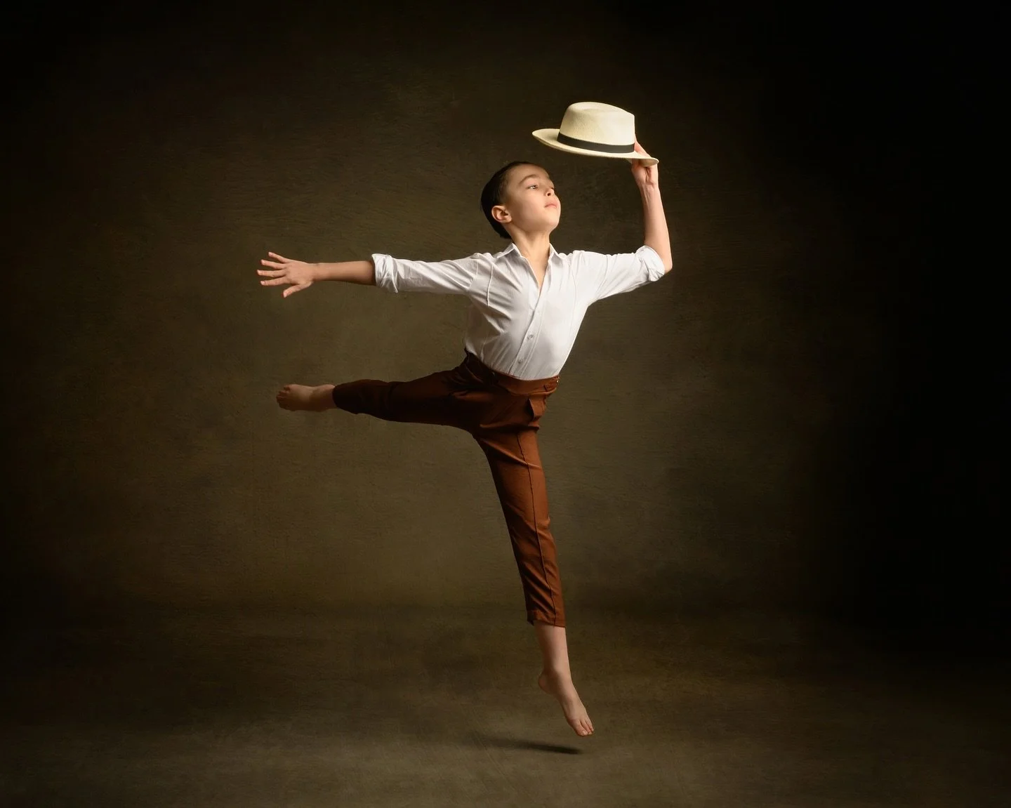 A young woman in a white shirt and brown pants balancing on one leg while jumping in the air, holding a hat with her hand.