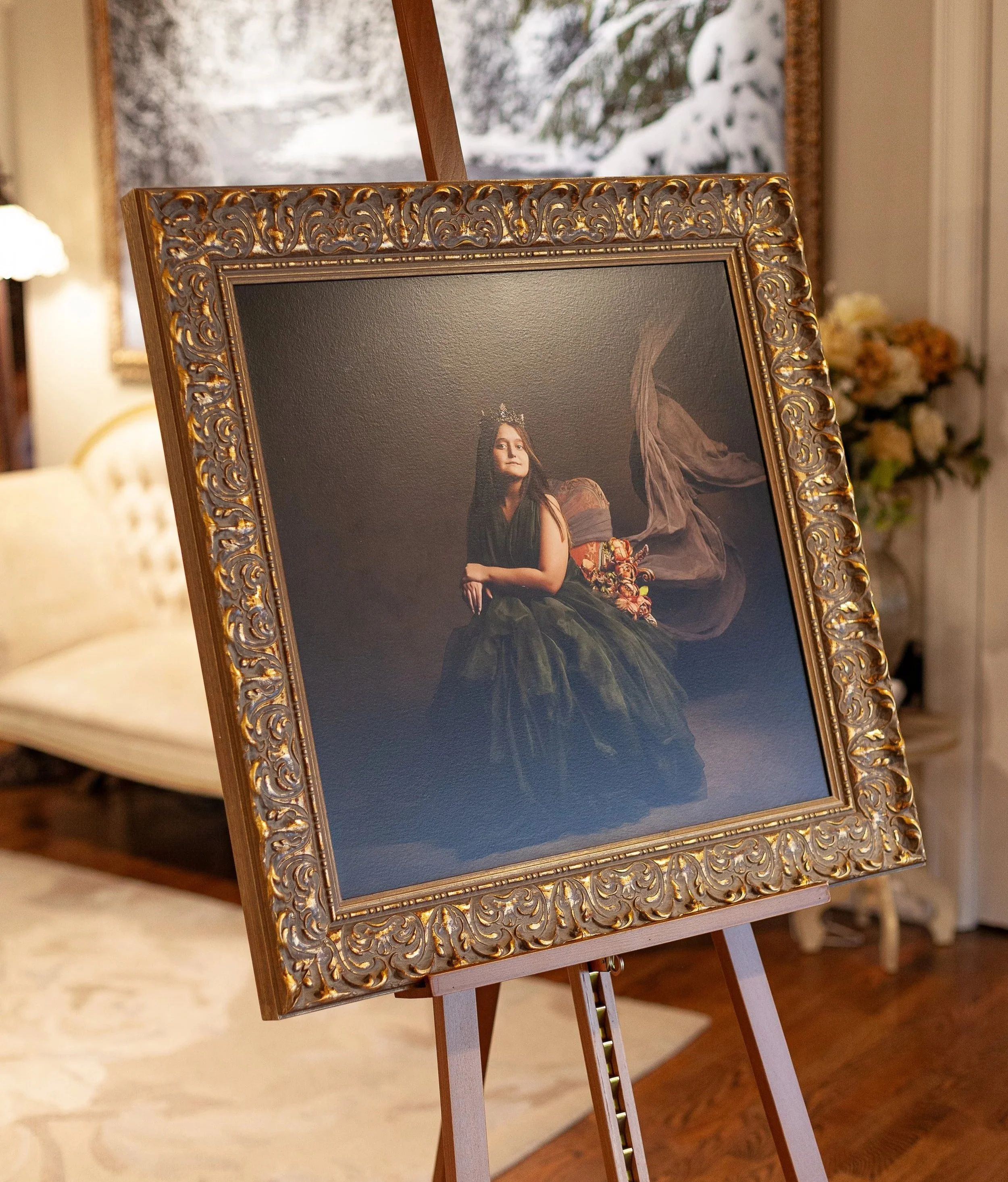 A framed portrait of a woman wearing a dark gown and a crown, sitting with her hands folded in her lap, depicted in a classical painting style, on an easel in a living room with a window showing snow outside.