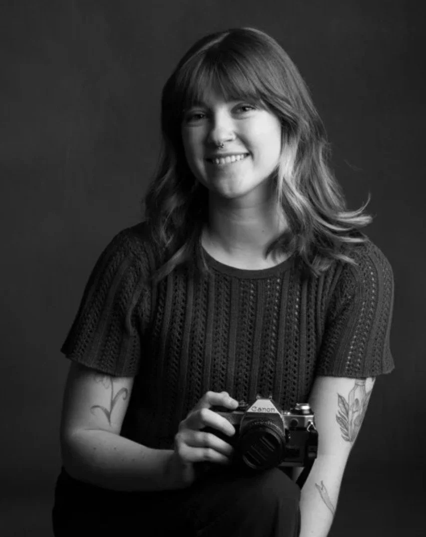 A young woman with shoulder-length wavy hair, smiling, holding a Canon camera, wearing a black knit top, with tattoos on her arms, posing against a dark background in a black and white photograph.