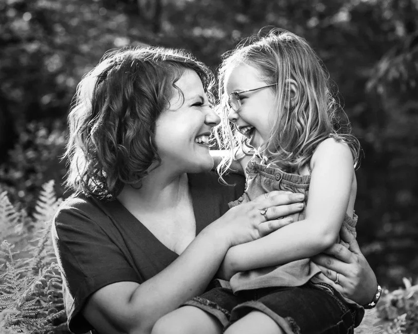 A woman and a young girl are smiling and laughing while looking at each other, outdoors with foliage in the background.