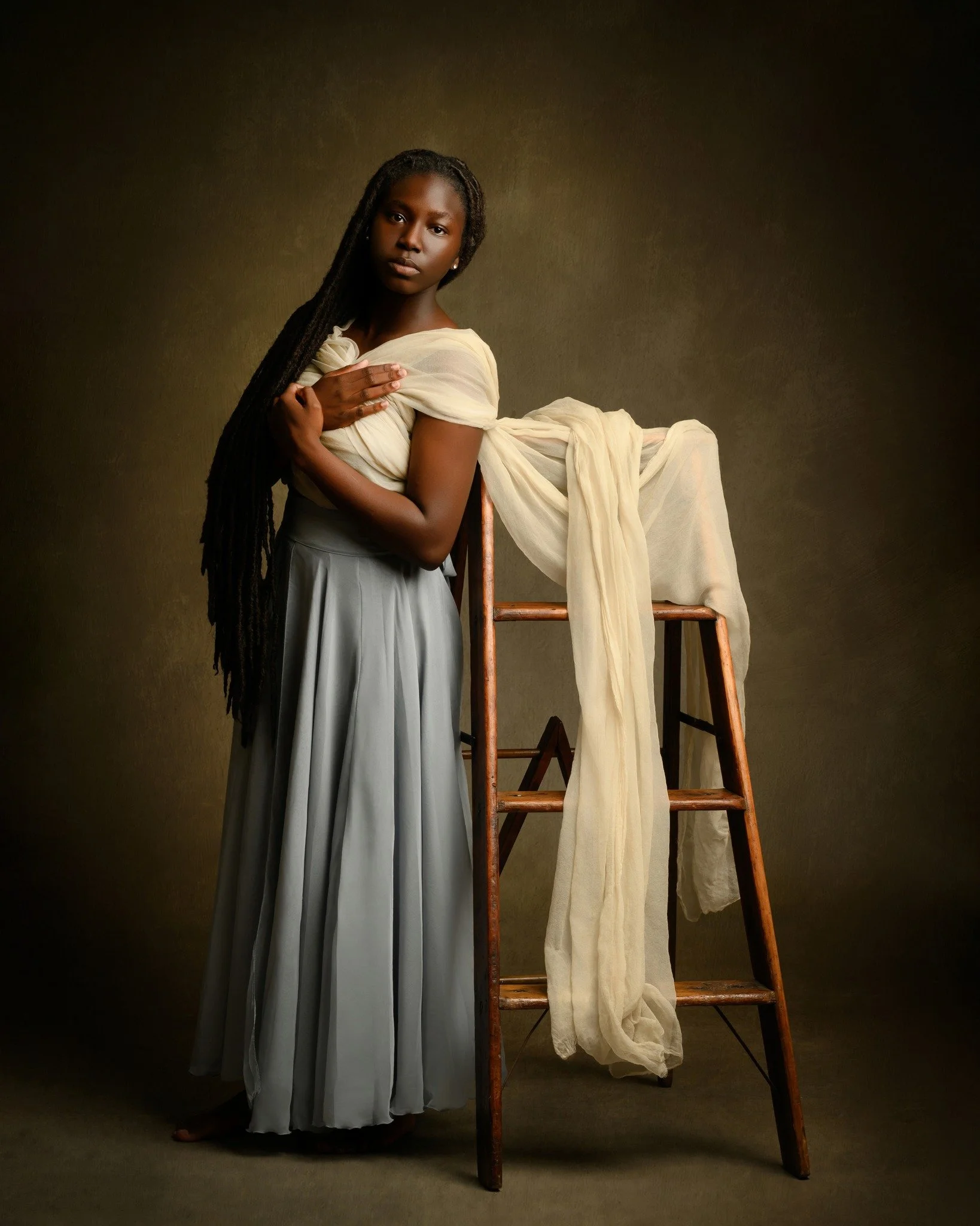A young woman with long dreadlocks wearing a light-colored dress, standing with one hand on her chest and the other resting on her waist, beside a wooden chair with a light-colored fabric draped over it, against a plain, dark background.