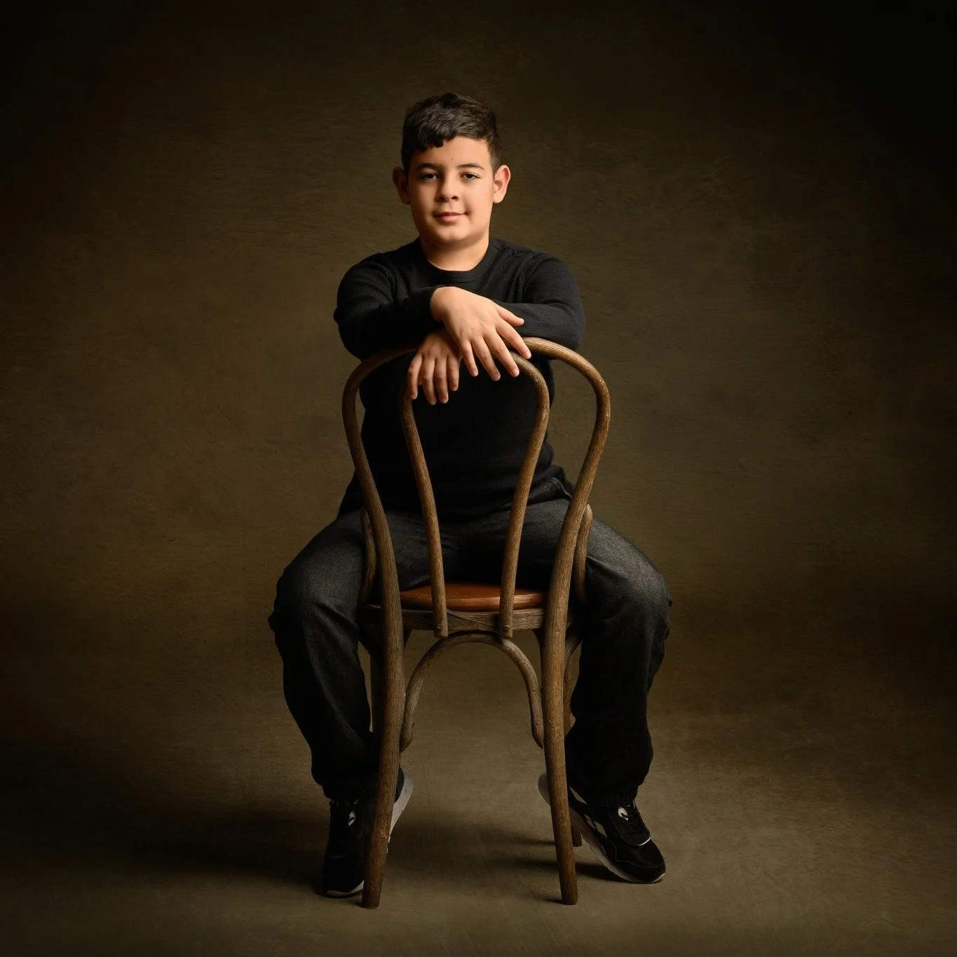 A young boy in black clothing sitting on a wooden chair, with his arms resting on the back of the chair, in a studio setting with a dark background.