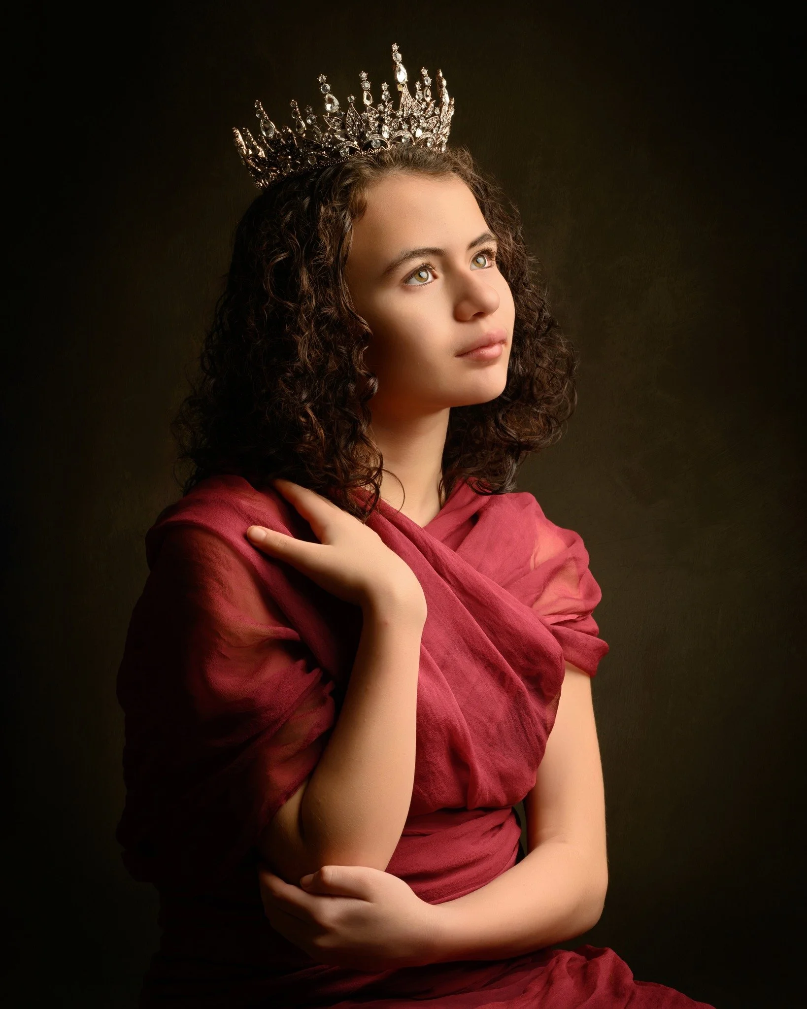 Young woman with curly hair wearing a tiara and red shawl, gazing thoughtfully.