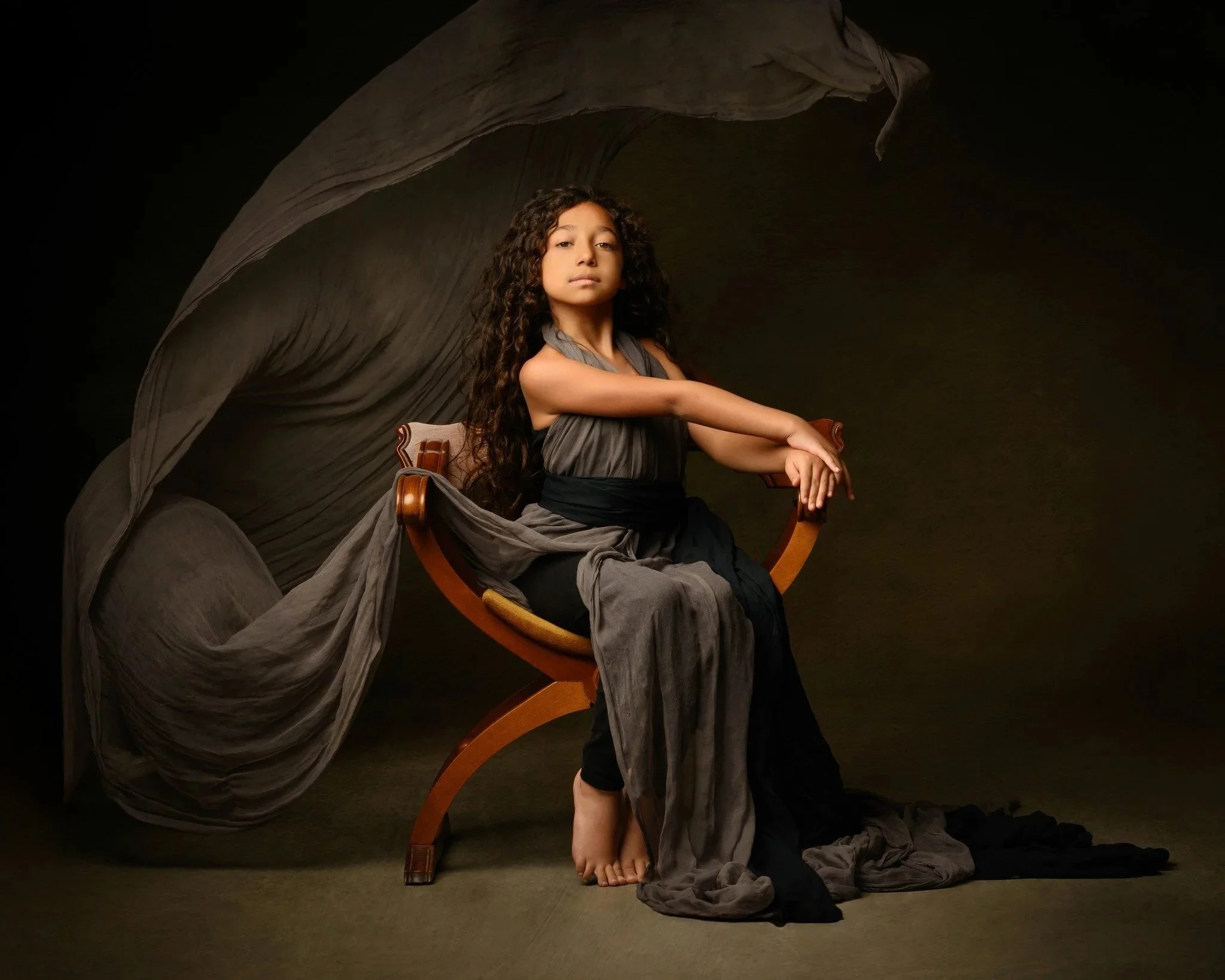 A woman with long, curly dark hair seated on a wooden chair in front of a dark background. She is wearing a gray dress with black accents and has an expressionless face. Her right arm rests on the armrest of the chair, and her left arm is crossed over her lap, with her bare foot visible on the floor. Grey fabric drapes around her, creating a flowing backdrop behind her.