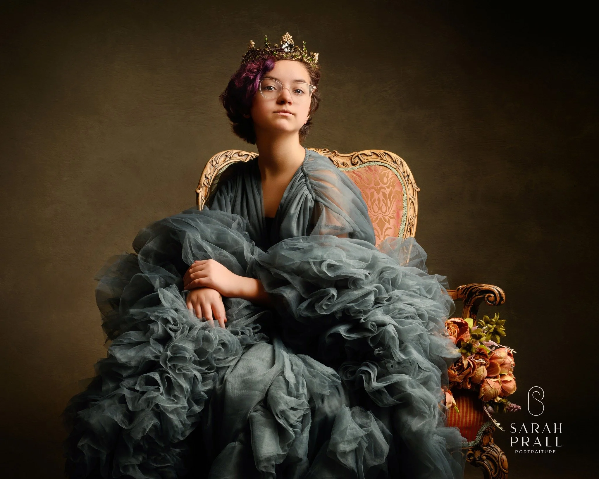 A young woman with glasses and purple-tinted hair, wearing a crown, seated on an ornate antique chair, dressed in an elaborate flowing gown with ruffled layers, against a dark background.