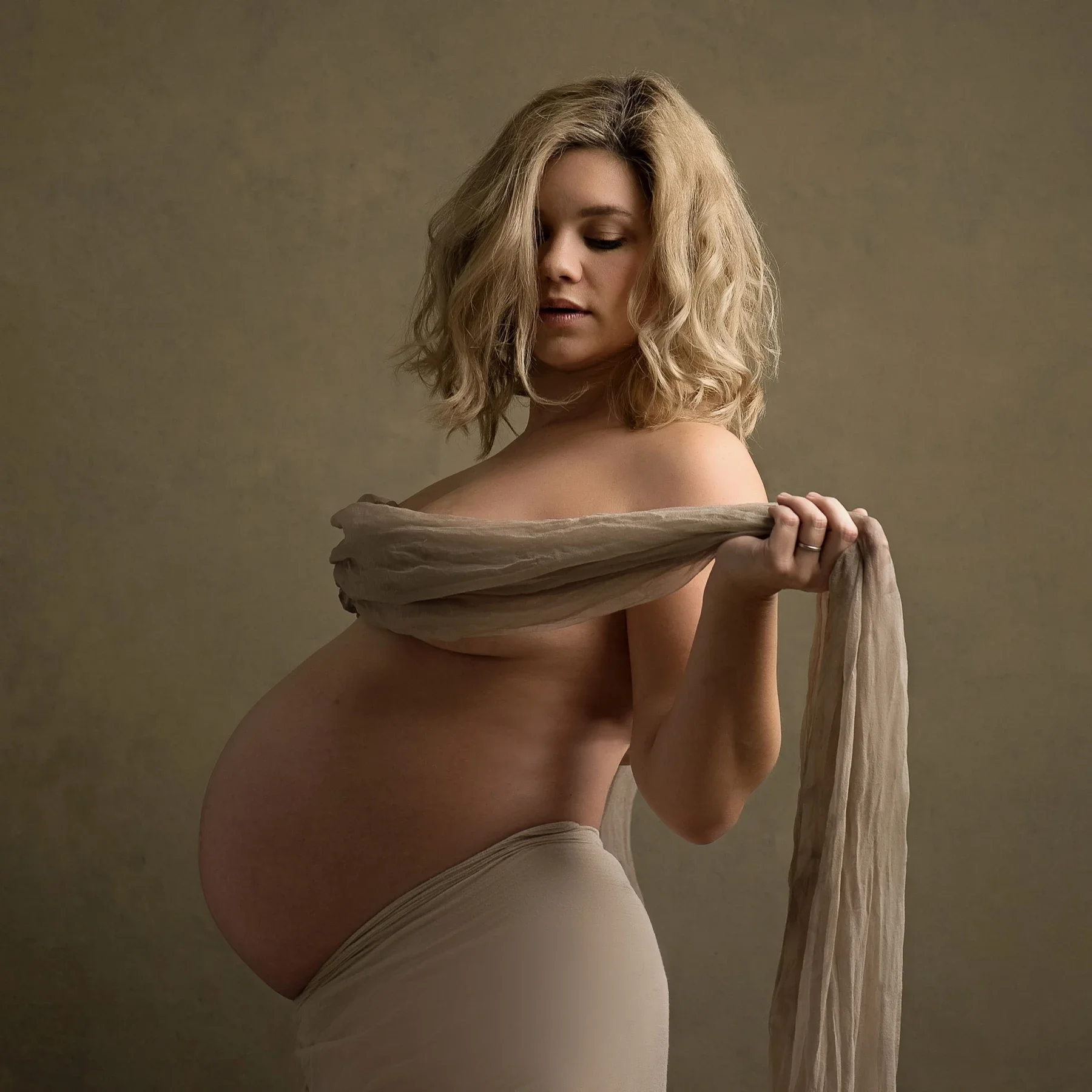 An expectant mother looking down, softly wrapped in taupe fabric