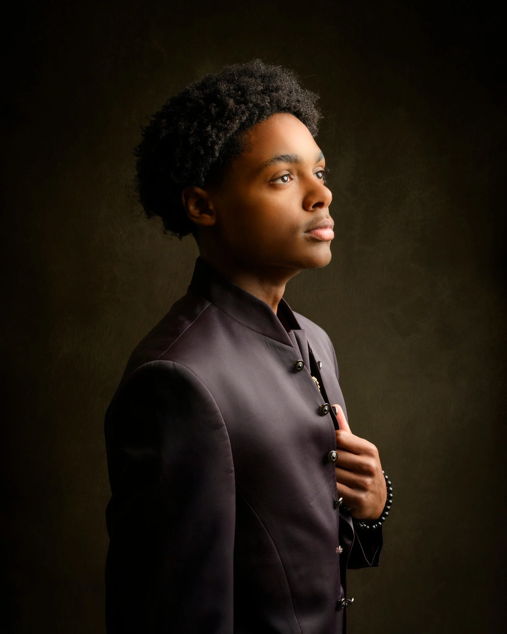 Portrait of a young woman with natural curly hair, wearing a dark satin suit, standing against a dark background, gazing thoughtfully into the distance.