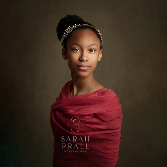 Portrait of a young African American girl with hair in a bun, wearing a decorative headband and a red shawl, against a plain dark background, promoting Sarah Prall portraiture.