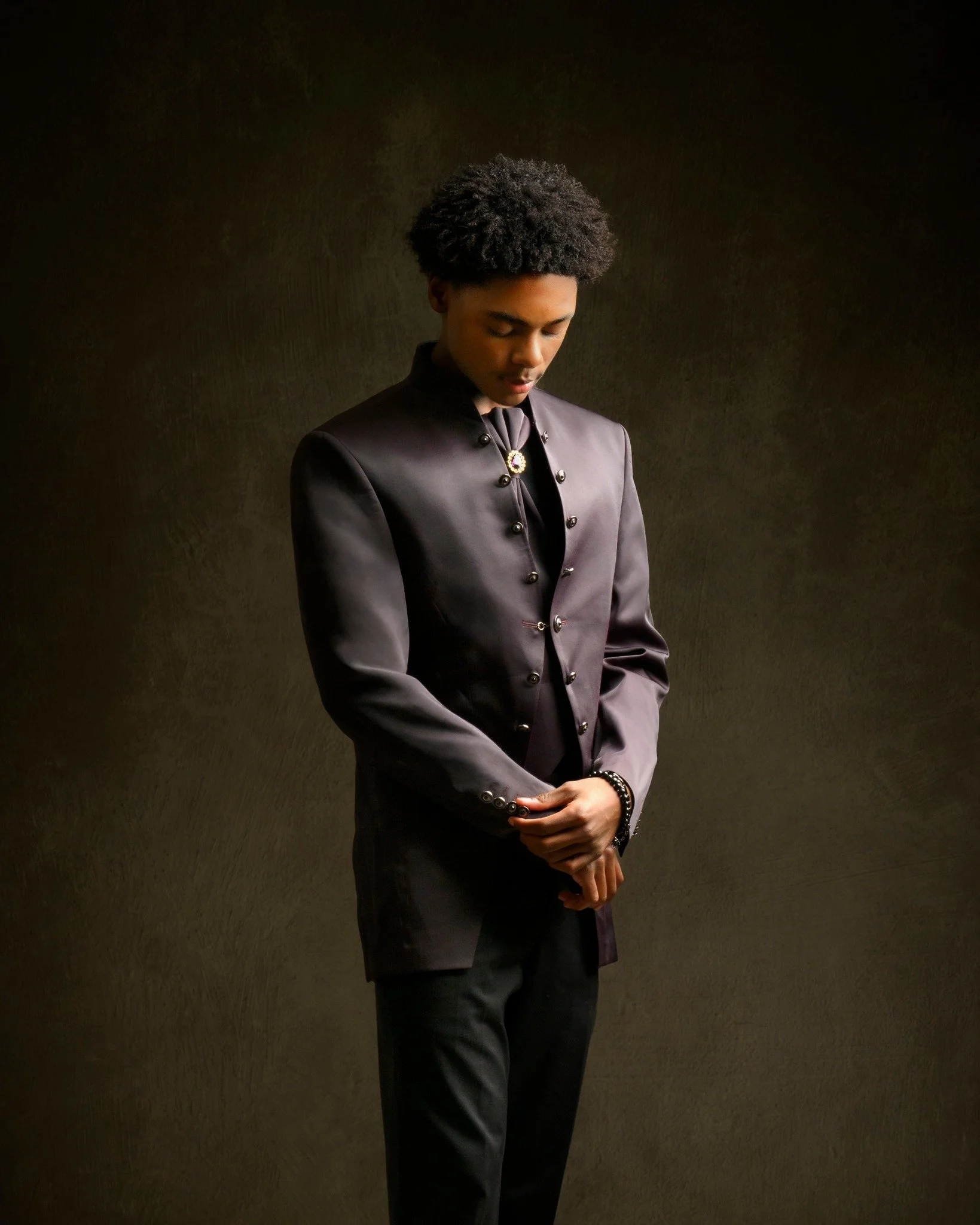 A young man in a dark, shiny, buttoned jacket with a high collar, standing against a dark, textured background, looking down with hands clasped.