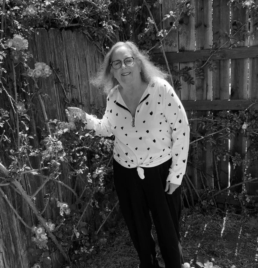 A woman with glasses and curly hair standing in a garden next to a wooden fence with flowering bushes, wearing a heart patterns shirt and dark pants.