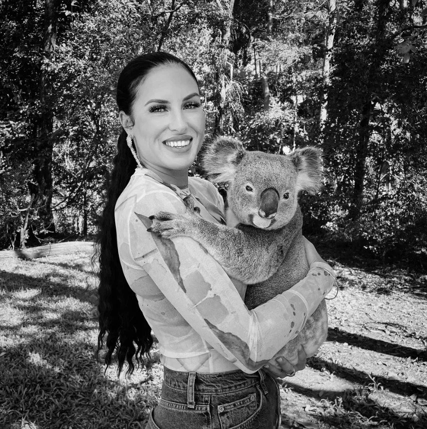 A woman with long dark hair holding a koala bear in a forest setting.