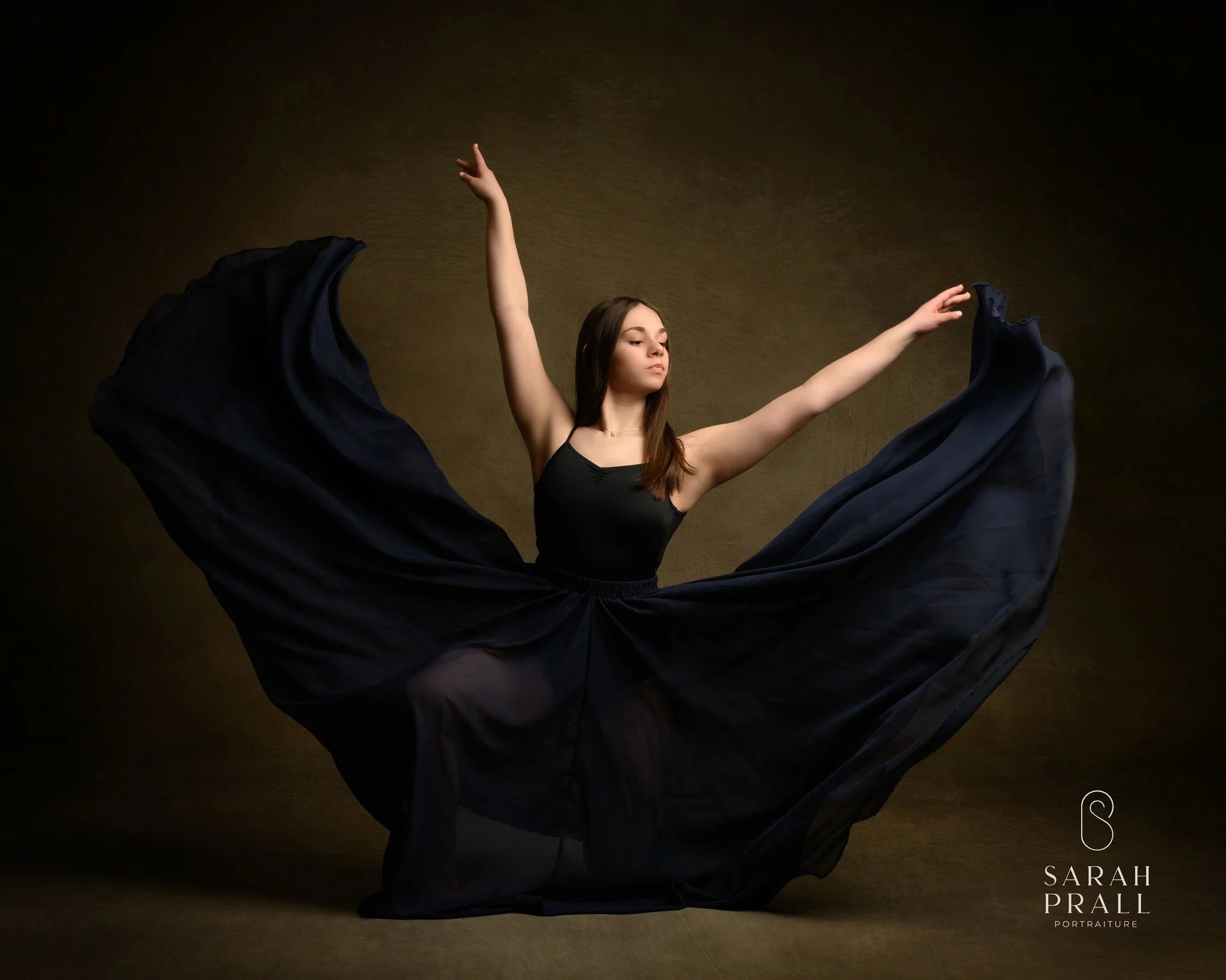 A young woman in a black dress dancing, with her arms raised and flowing black fabric creating a wide semi-circle around her. The background is dark brown.