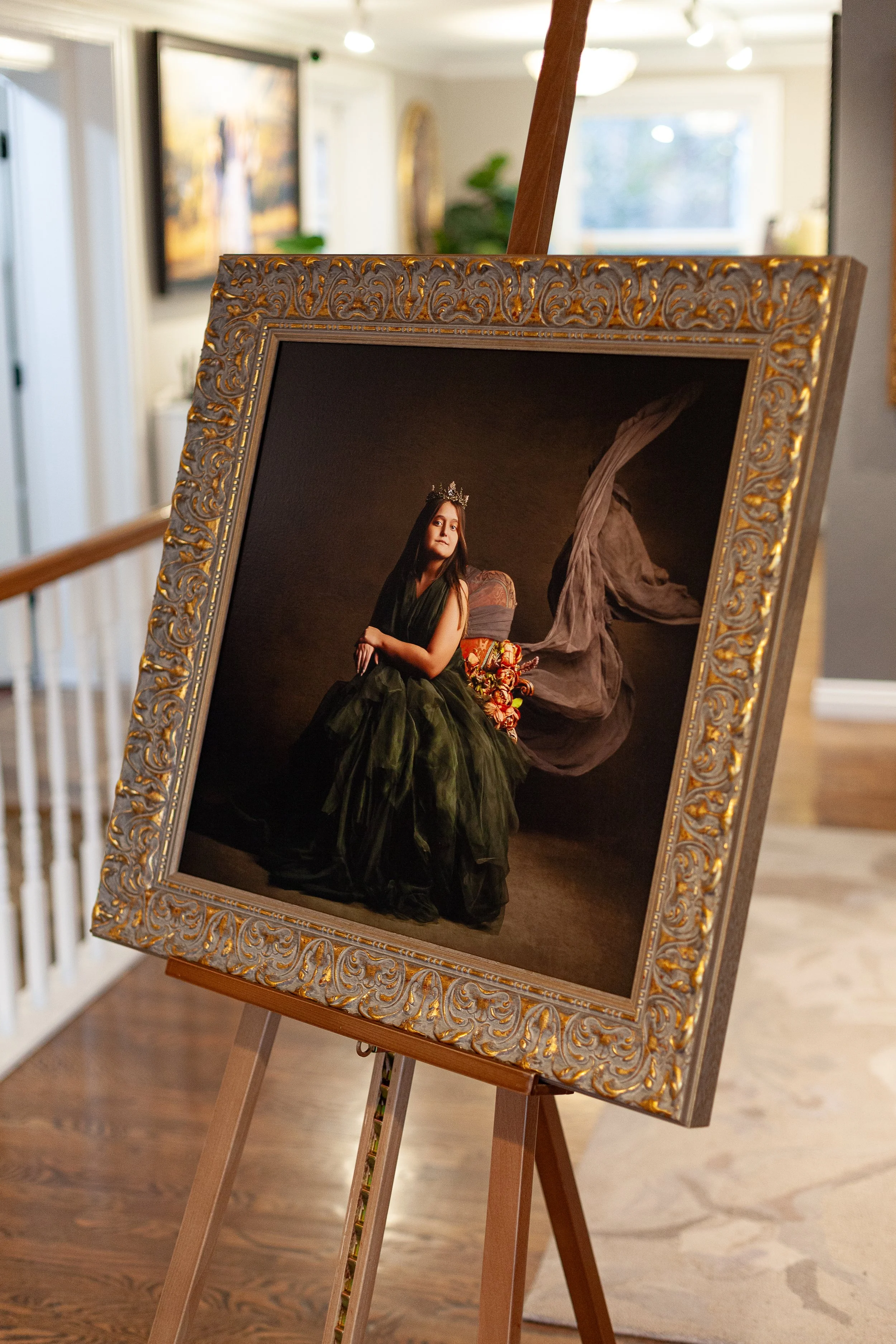 A portrait of a woman sitting on a chair, wearing a dark green dress and a crown, with floral arrangements behind her, displayed in an ornate gold frame on an easel inside an art gallery.
