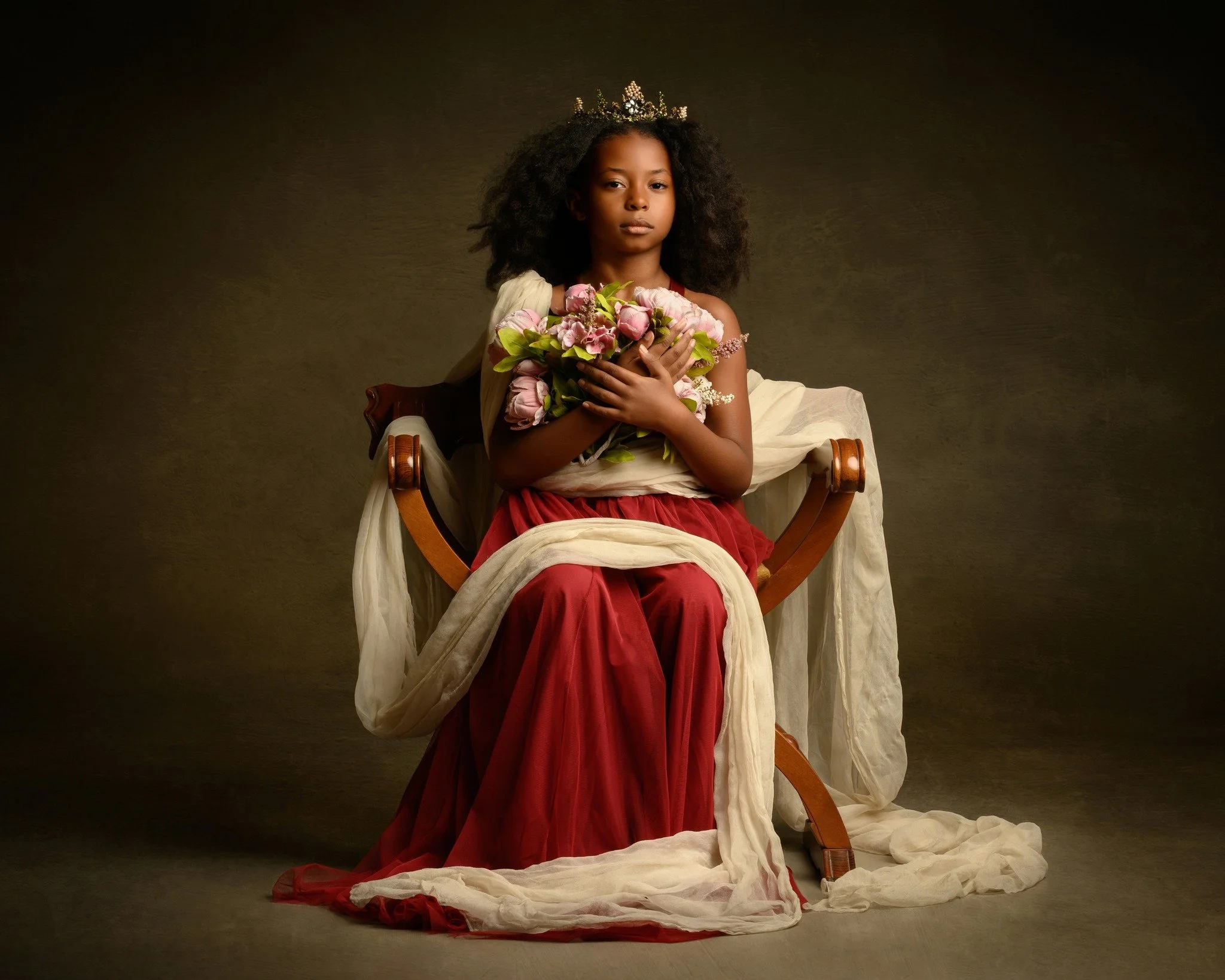 A young girl with curly hair and a tiara, dressed in a royal-like gown, sitting on a chair holding a bouquet of pink flowers, with an elegant, vintage-style backdrop.
