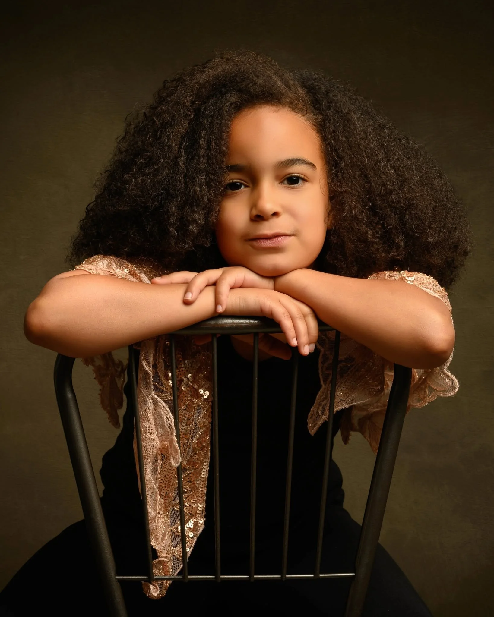 A young girl with curly hair resting her chin on her folded arms, sitting behind and gripping the backrest of a black chair. She is wearing a lace and sequined dress and looking directly at the camera with a calm expression, against a plain dark background.