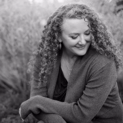 Black and white portrait of a woman with curly hair smiling and looking down, outdoors background.