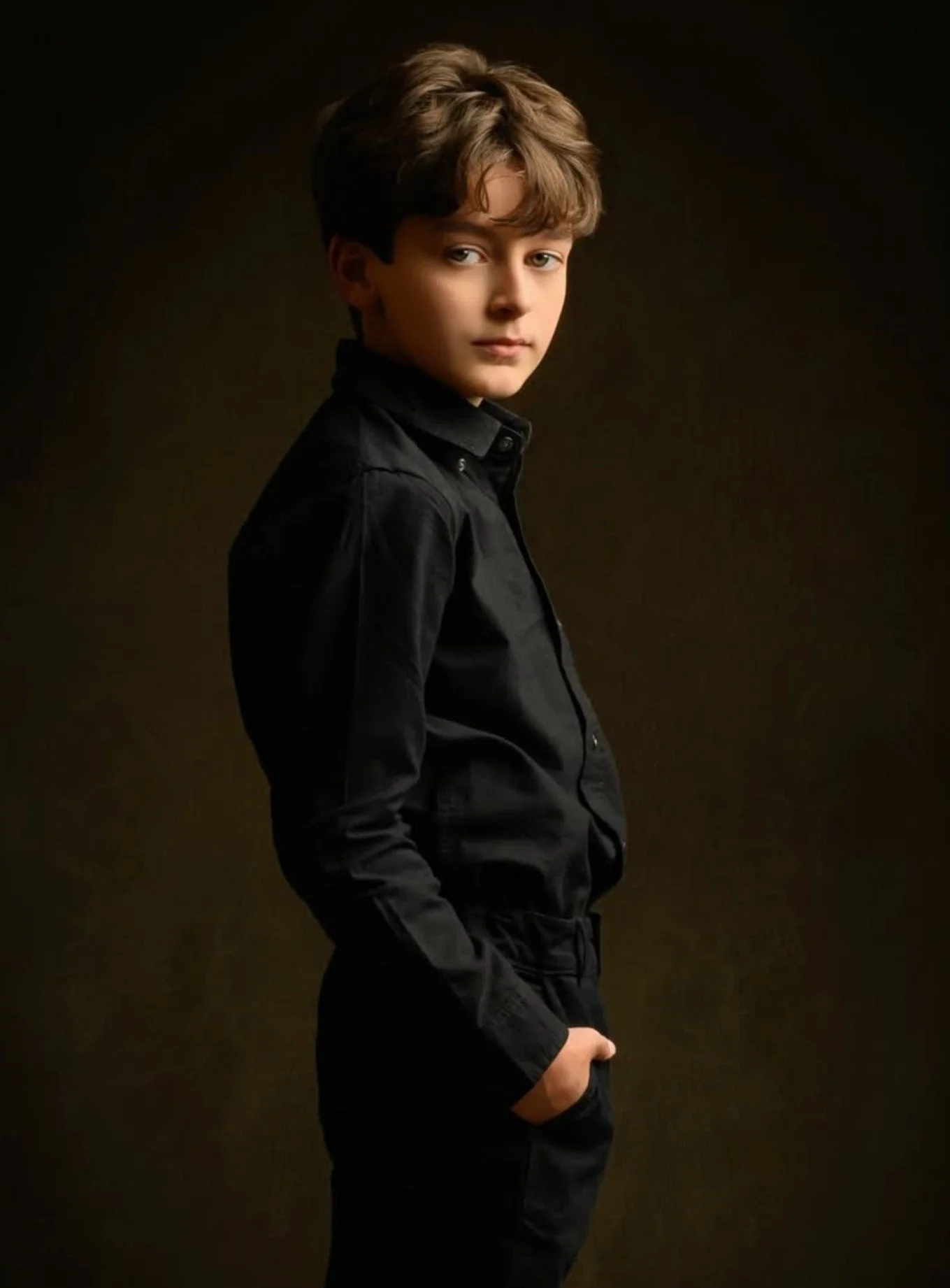 A young boy with brown hair and light skin wearing a black button-up shirt, standing with his right hand in his pocket against a dark background.
