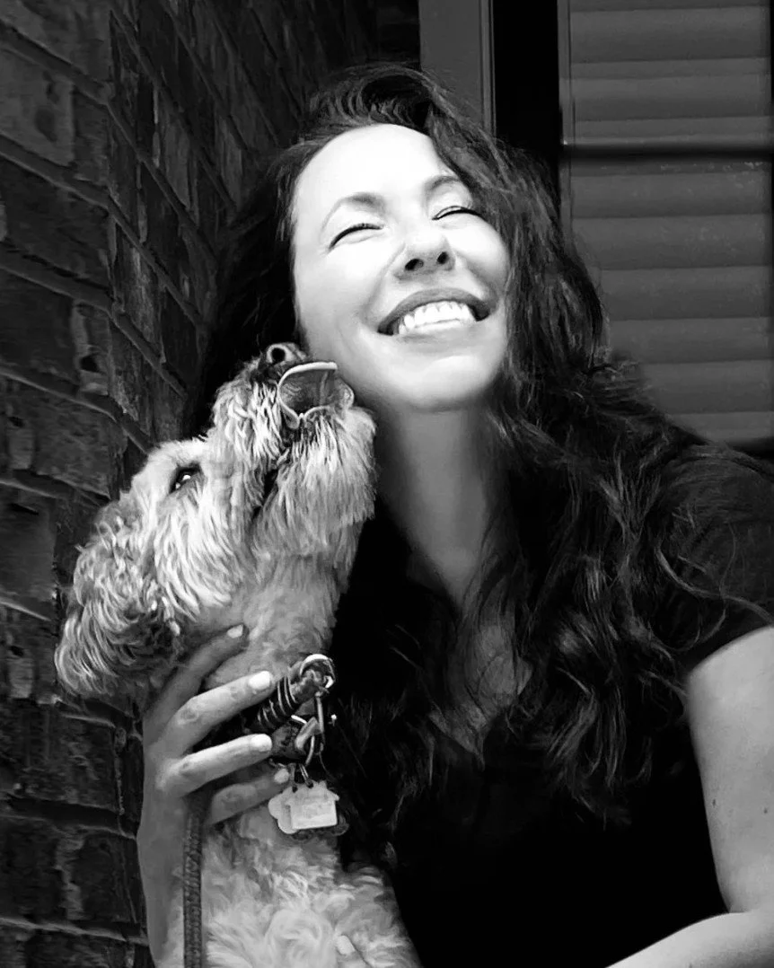 A woman with long curly hair smiling and hugging a dog, both in black and white.