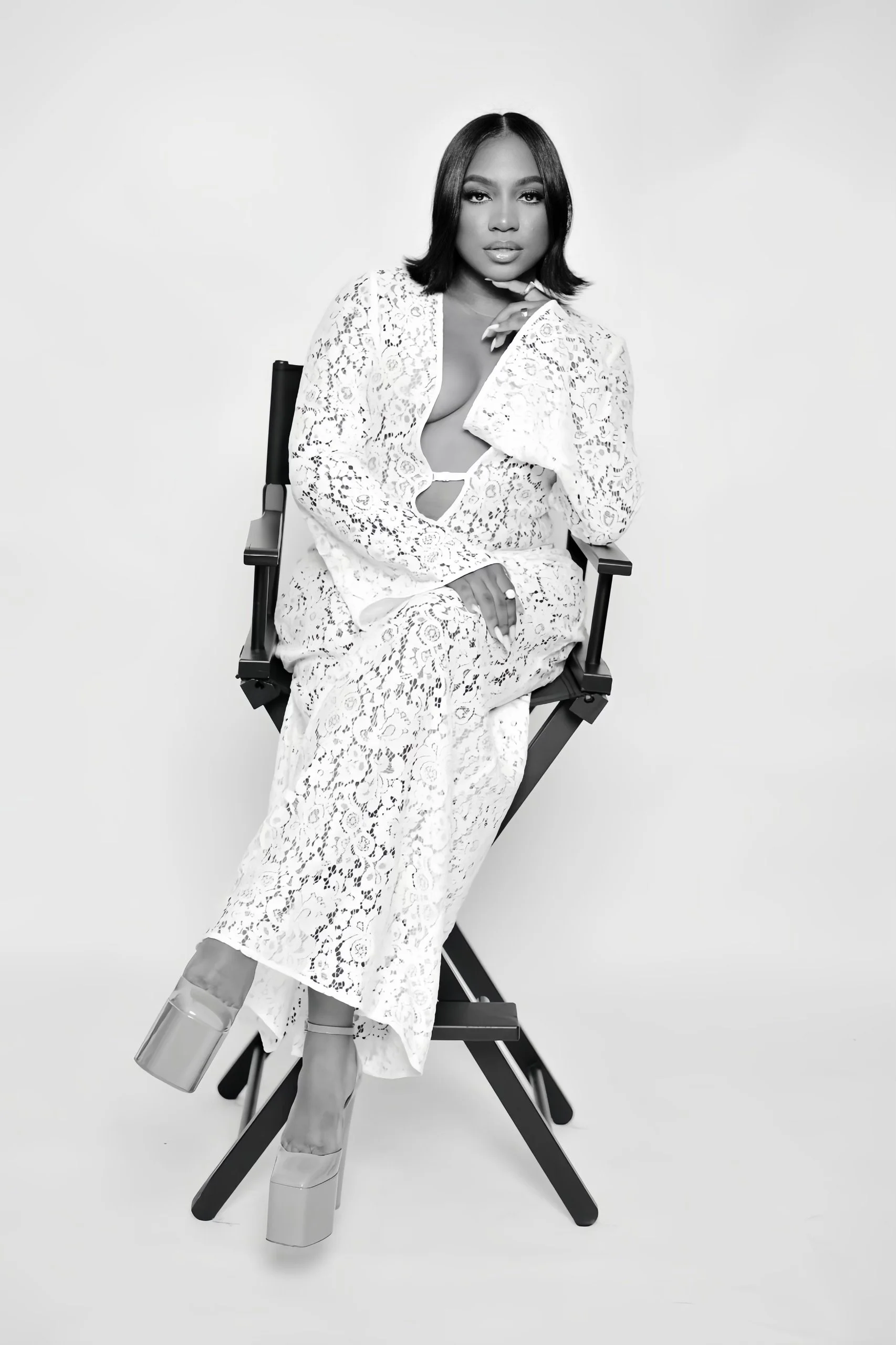 Black and white photo of a woman with dark, shoulder-length hair sitting on a director's chair, wearing a white lace outfit and high platform shoes, posing confidently with her hand near her face.