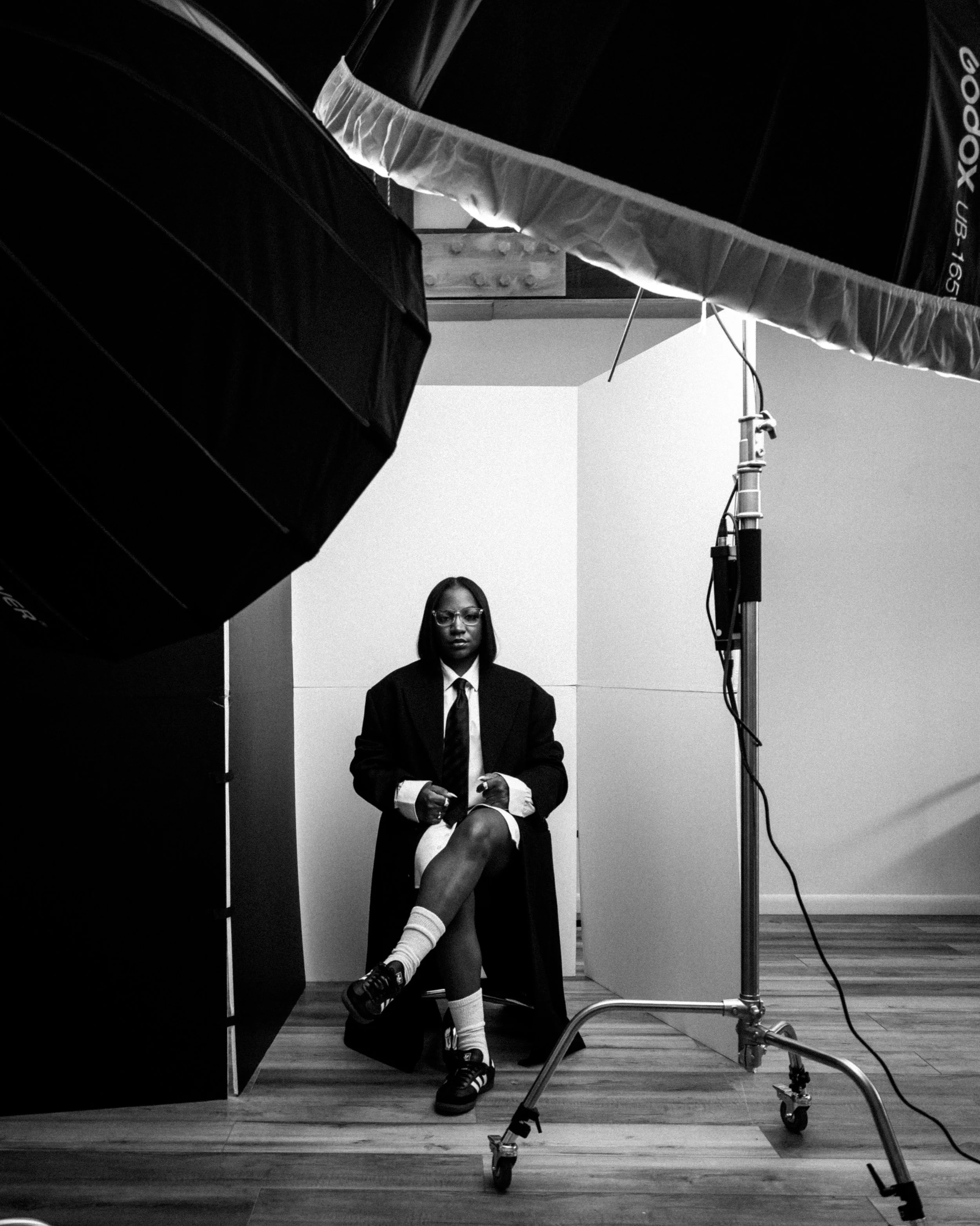 A woman sitting in a photography studio, wearing a suit, glasses, and sneakers, with a backdrop and studio lighting equipment.