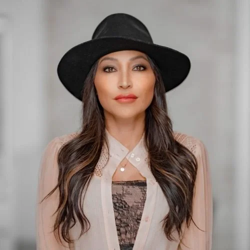 A woman with long dark hair wearing a black hat and a beige blouse, standing in front of a blurred neutral background.