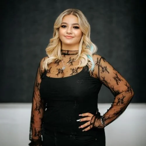 Young woman with blonde, wavy hair and a slight smile, wearing a black sheer lace top with floral patterns and a black tank top underneath, posing against a dark background.