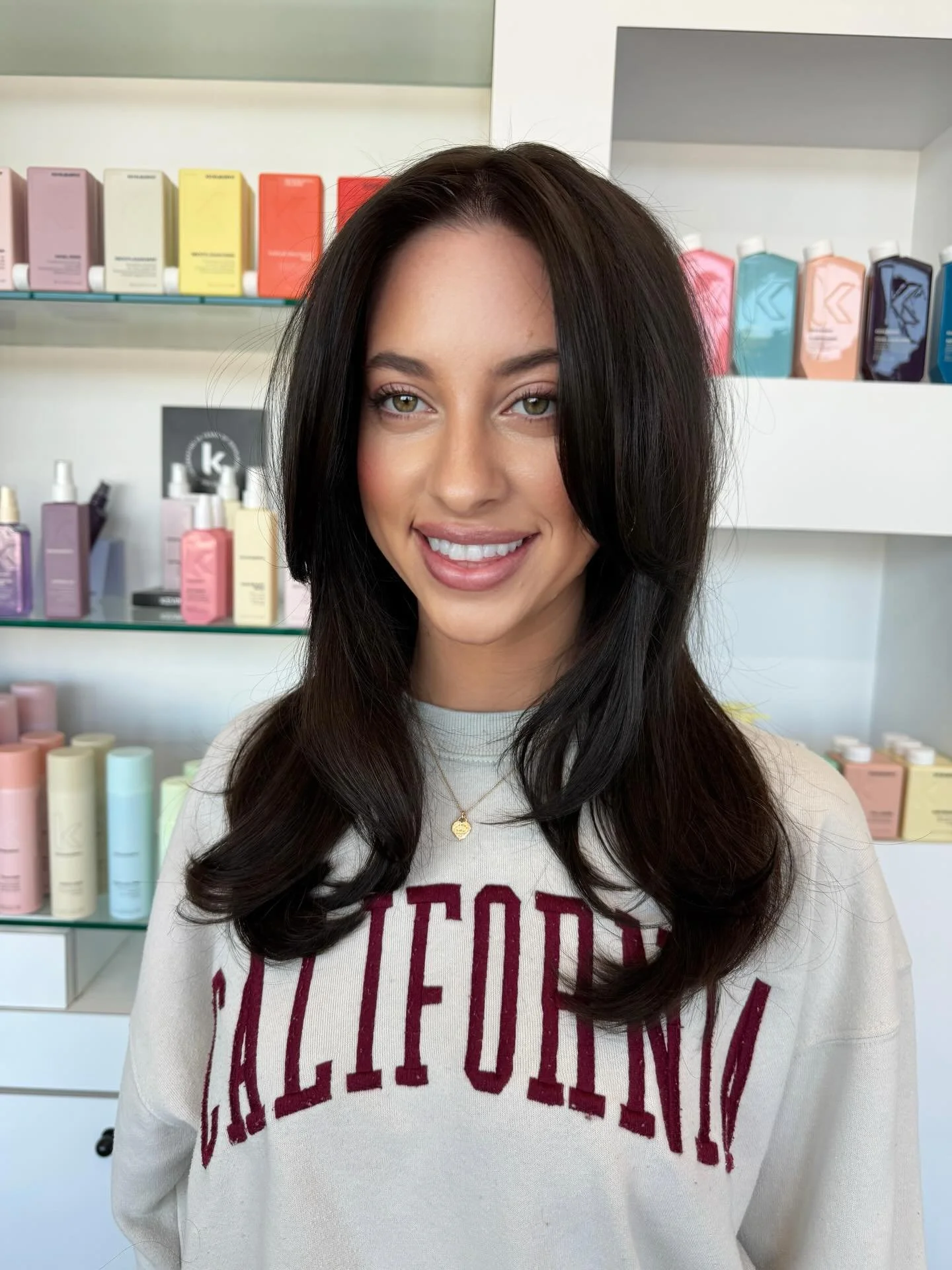 A young woman with dark brown hair and light skin smiling, wearing a beige sweatshirt with red text, standing in front of shelves with colorful bottles and boxes.