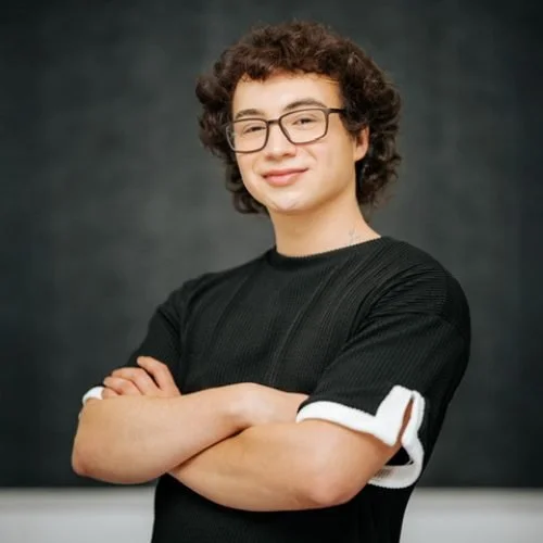 Young man with curly hair and glasses, wearing a black shirt, standing with arms crossed and smiling.