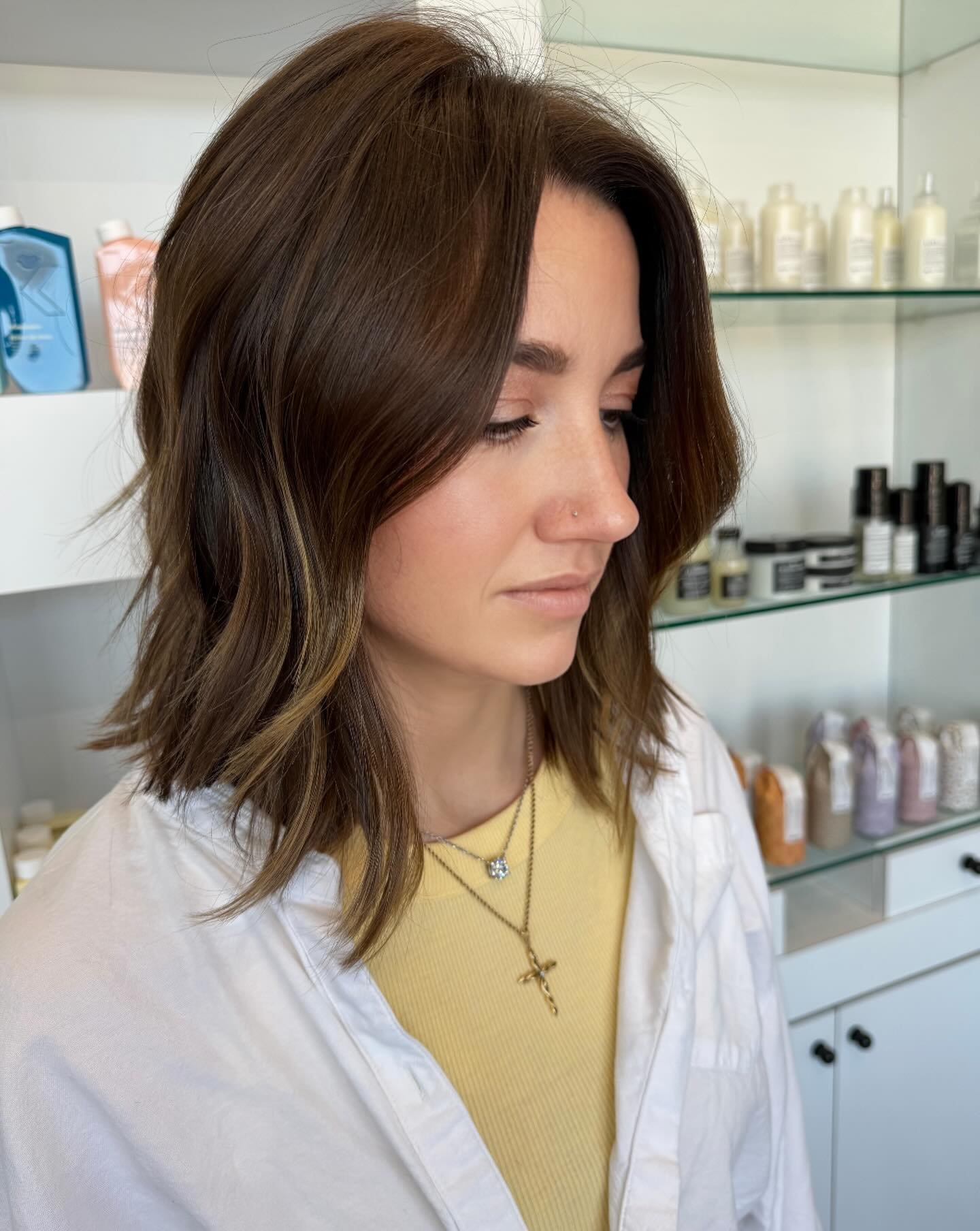 Young woman with shoulder-length brown hair, wearing a yellow top and layered necklaces, looking downward in a medical or beauty clinic with shelves of skincare products in the background.