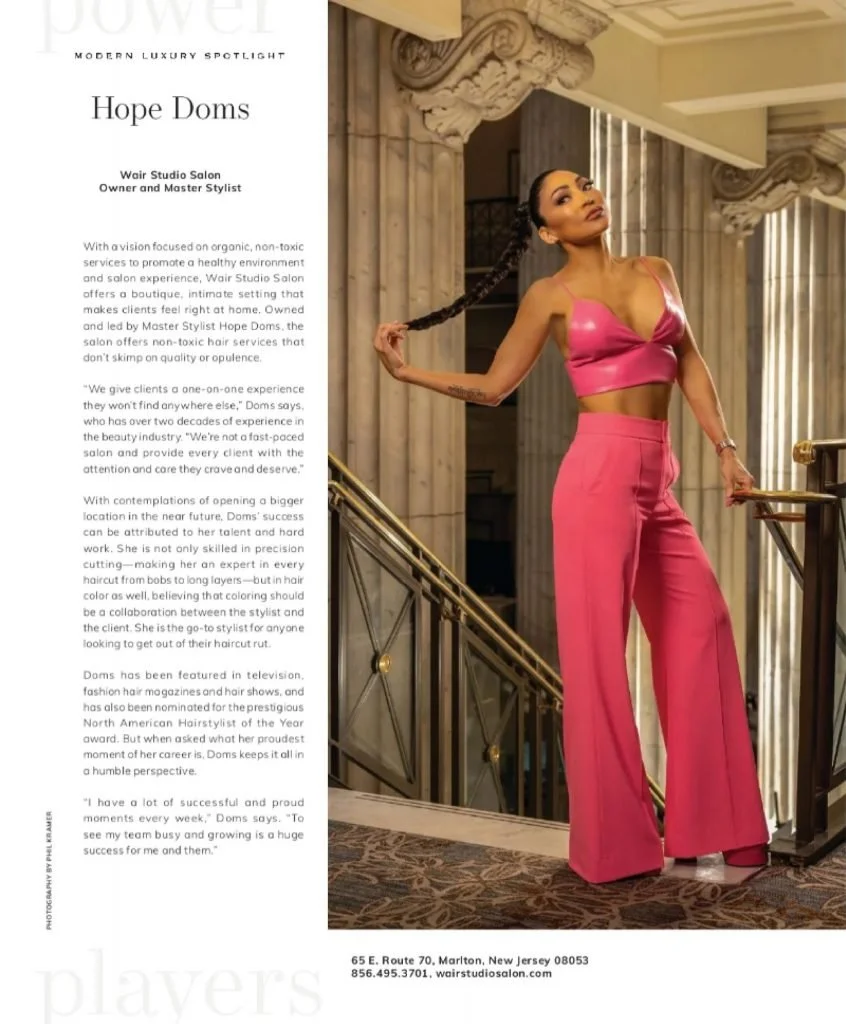 A woman in a pink satin two-piece outfit with a crop top and wide-leg pants poses on a staircase in a luxurious building with ornate architecture and beige marble columns.