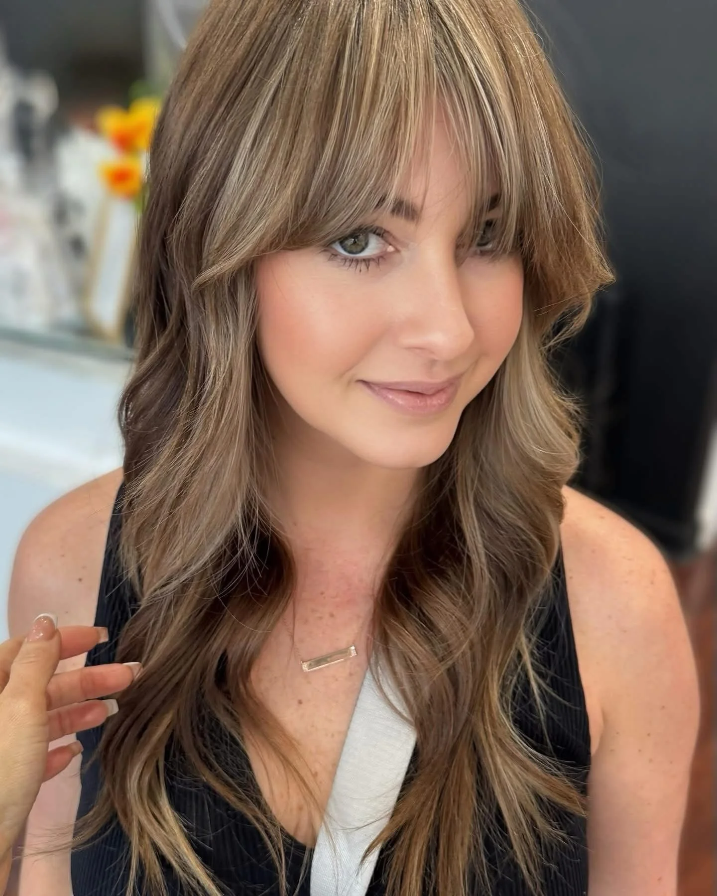 A woman with wavy, light brown hair and highlights, smiling softly at the camera, wearing a black sleeveless top and a delicate gold necklace.