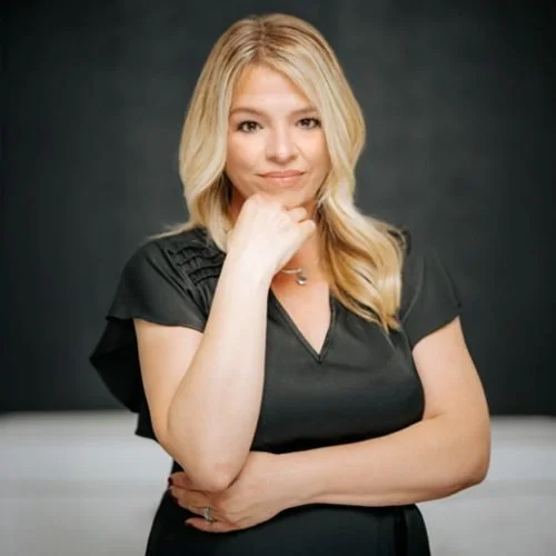A woman with blonde hair and fair skin, wearing a black top, standing with her arms crossed and resting her chin on her hand, against a black background.