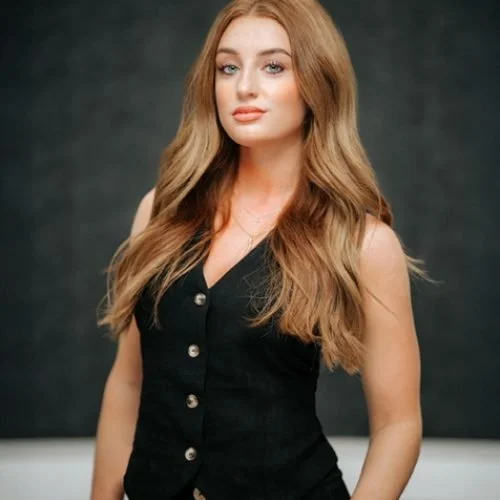 Young woman with long, wavy blonde hair wearing a black sleeveless button-up top, standing against a dark background