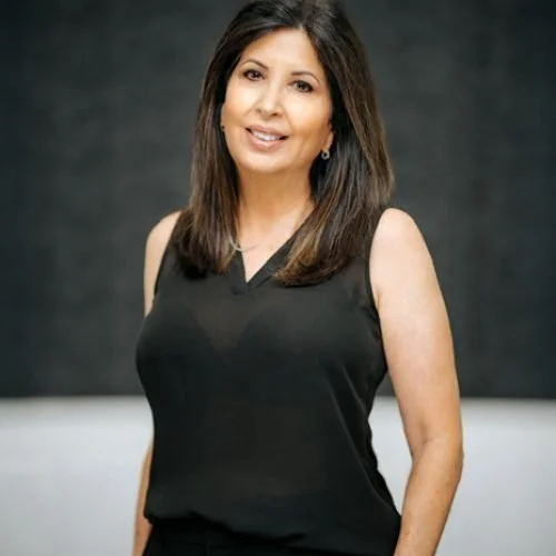 A woman with shoulder-length dark hair wearing a sleeveless black top standing in front of a dark gray background.