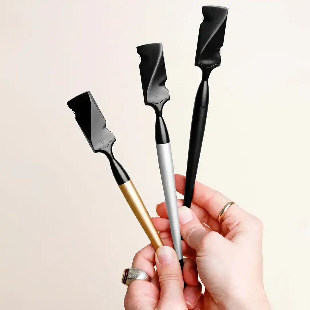 A hand holding three black spatulas with different colored handles: gold, silver, and black, against a plain white background.