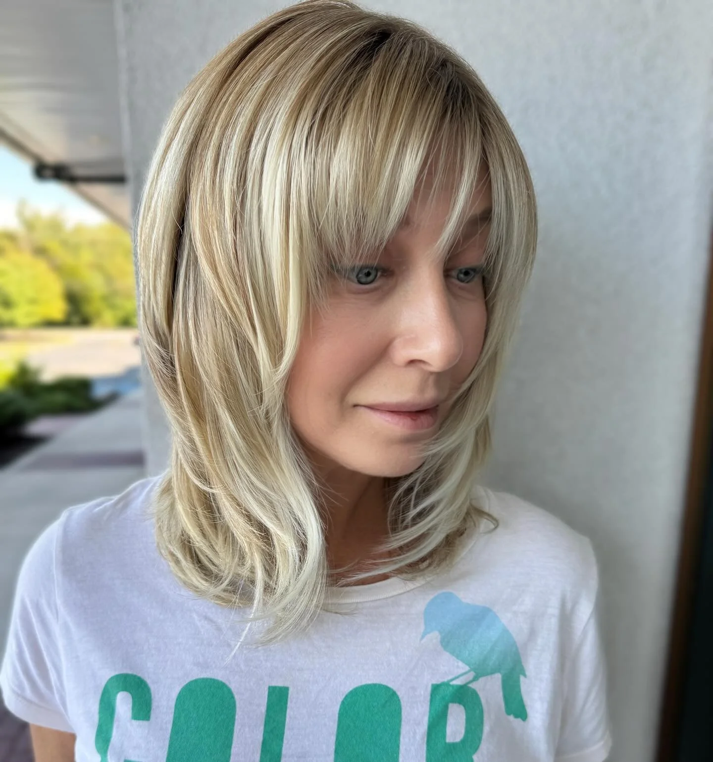 A woman with blonde hair and blue eyes wearing a white T-shirt with a blue bird graphic standing outside near a wall.