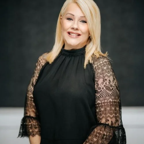 A young woman with blonde hair smiling, wearing a black dress with lace sleeves, standing against a dark background.