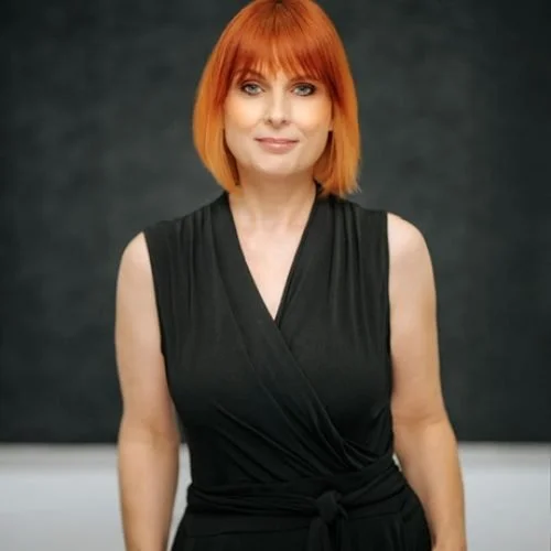 A woman with shoulder-length red hair and bangs, wearing a sleeveless black dress, standing against a dark background.