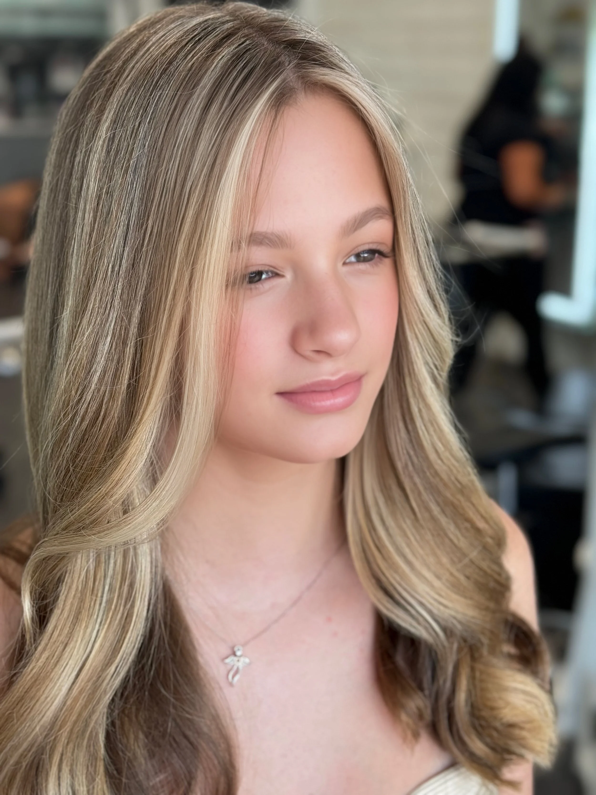 A young woman with long blonde hair and a light pink complexion, smiling softly, wearing a necklace, in an indoor setting.